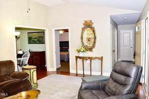 A living room with tan walls, leather recliners, a side table, and a console table topped with a flower arrangement beneath an ornate mirror. Doorways lead to a dining area, bedroom, and hallway.