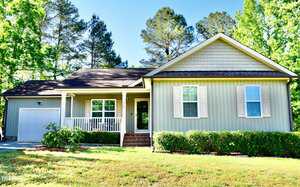 A single-story, light gray house with white trim and shutters at 1980 Ferbow Street in Creedmoor features a front porch, attached one-car garage, and manicured lawn with bushes. Tall trees surround the property under a clear blue sky.