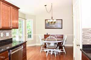 A bright kitchen dining area with a round wooden table, four white chairs, a sideboard, wood floors, and a window overlooking greenery. A chandelier hangs above the table and a framed picture is on the wall.