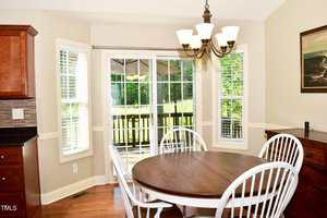 A dining area with a round wooden table and four white chairs is shown next to windows and a glass door leading to a deck outside. A chandelier hangs above the table, and sunlight fills the room.