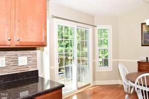 A kitchen with wood cabinets, black countertops, and a tile backsplash. A sliding glass door opens to a deck outside. A round wooden table with white chairs sits near a window with white blinds.