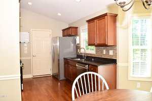 A kitchen with wooden floors, brown cabinets, stainless steel appliances, a black countertop, a tiled backsplash, and a round wooden dining table with white chairs near a window with blinds.