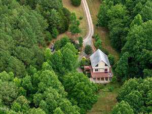 Aerial view of a two-story house with a porch, surrounded by dense green trees and a grassy yard. A gravel driveway curves around the property, leading to a parked vehicle partially visible in the trees.