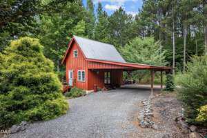A small red two-story cabin with a metal roof and a covered carport sits among trees and greenery, with a gravel driveway and rock-edged landscaping in front.