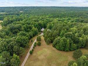 A house is partially visible through dense, green trees, surrounded by a large forested area. A long gravel driveway leads to the house, bordered by open grassy fields and clusters of mature trees.