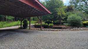 A gravel driveway curves around a covered carport with a wooden roof. There are landscaped garden beds with rocks and various trees and shrubs in the background. The scene is outdoors with natural greenery.