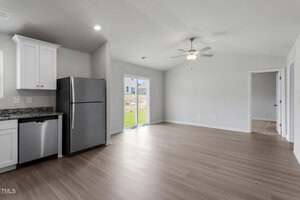 A modern, unfurnished kitchen and living area with light wood floors, white cabinets, stainless steel appliances, a ceiling fan, sliding glass doors, and an adjacent room visible through an open doorway.