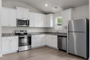 Modern kitchen with white cabinets, stainless steel appliances including a refrigerator, dishwasher, oven, and microwave, granite countertops, and light-colored wood flooring. A small window above the sink lets in natural light.