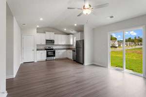 Open-plan kitchen and living area with white cabinets, stainless steel appliances, light wood flooring, ceiling fan, and large sliding glass doors overlooking a grassy suburban street with houses and blue sky.