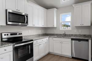 Modern kitchen with white cabinets, stainless steel appliances including a stove, microwave, and dishwasher, granite countertops, a single window above the sink, and light wood flooring.