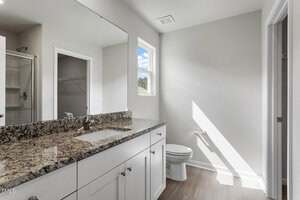 A modern bathroom with a granite countertop, single sink, white cabinets, a toilet, and a window letting in natural light. A shower with glass door and a walk-in closet are visible in the background.