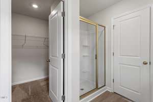 A small shower with gold trim is next to two white doors; one door opens to a carpeted walk-in closet with wire shelving, and the other door is closed. The shower area has light-colored walls and flooring.