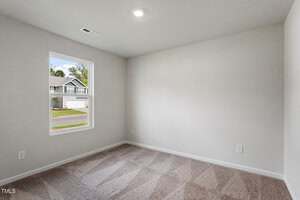 Empty room with light gray walls, beige carpet, a single window showing a suburban street, white baseboards, ceiling light, and electrical outlets visible on two walls.