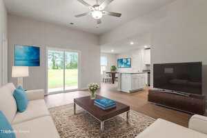 A modern living room with a white sofa, a wooden coffee table, a flat-screen TV, and a patterned rug. Sliding glass doors open to a green lawn. The room connects to a white kitchen with bar seating.
