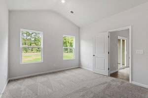 A bright, empty room with light-colored walls, two windows showing greenery outside, beige carpet flooring, a white ceiling, and an open door leading to a hallway with wood-like flooring.