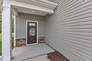 A covered entryway with gray vinyl siding, a black door with a glass window, a brick accent along the bottom, and a concrete porch. There is a small section of mulch and some grass visible to the left.