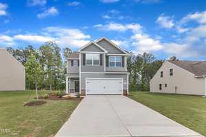 A two-story suburban house with gray siding and white trim, featuring a double garage and a wide concrete driveway. The front yard has small landscaping and is surrounded by grassy lawns and neighboring houses.
