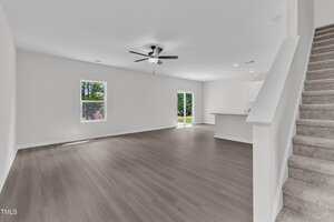 Open-concept living area with light-colored walls and wood flooring, featuring a ceiling fan, two windows, sliding glass doors, a kitchen with a breakfast bar, and carpeted stairs leading to the upper floor.