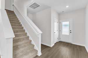A bright entryway with light-colored walls, wood-style flooring, a white front door with frosted glass, a closet door, and a carpeted staircase with a white railing leading to the upper floor.