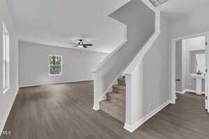 A modern, empty living room with light gray walls, wood-look flooring, a ceiling fan, and a carpeted staircase leading up. A small bathroom with a pedestal sink and mirror is visible to the right.