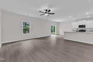 A spacious, empty open-concept living area with light wood flooring, white walls, a ceiling fan with lights, two windows, a glass door, and a modern kitchen featuring white cabinets and stainless steel appliances.