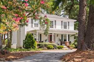 A two-story white house with black shutters and a covered front porch, surrounded by trees and greenery. A gravel driveway leads to the entrance, and flowering branches frame the view.