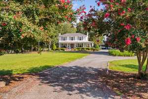 A two-story white house with a porch sits at the end of a gravel driveway on 3123 Brassfield Road in Creedmoor, surrounded by green lawn and trees with pink flowers under a clear blue sky.