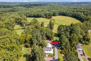 Aerial view of a rural property with a white house, red-roofed building, several trees, open green fields, a pond, and surrounding forested areas under a blue sky.
