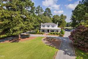 A two-story white house with a covered front porch sits near the center of a large, well-kept lawn surrounded by tall trees. A circular driveway leads to the front entrance, and several cars are parked in the background.