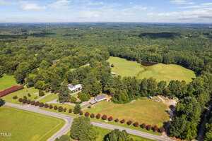 Aerial view of a rural property with several houses, open fields, fenced areas, a small pond, a tree-lined road, and dense forest surrounding the area under a partly cloudy sky.