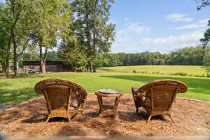 Two wicker chairs and a small table sit on a mulched area overlooking a green lawn and open field with trees and a barn in the background under a partly cloudy sky.