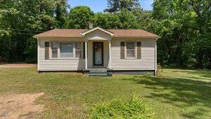 A small, single-story beige house with a brown roof, three front windows with shutters, a central door with a wreath, and a grassy yard, surrounded by trees in the background.