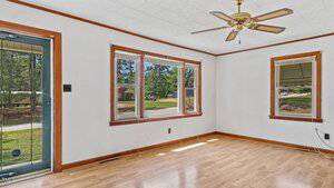 A bright, empty room with light wood flooring, white walls, three large windows, a ceiling fan with a light, and a glass door. Trees and a lawn are visible outside through the windows.