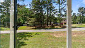 View from a porch showing a grassy yard, a paved road, several tall pine trees, and buildings including garages and houses with multiple vehicles parked outside under a sunny sky.