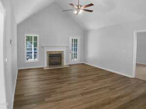 A spacious, empty living room with light gray walls, wood flooring, a white fireplace, a ceiling fan with lights, and two glass doors leading outside. A window is on the left, and an open doorway is on the right.