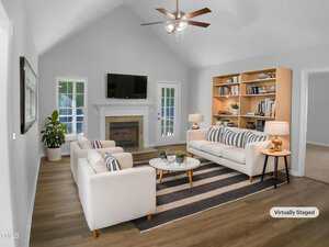 A living room with white walls, wood flooring, a fireplace, mounted TV, two white sofas, a striped rug, a coffee table, a wooden bookshelf, lamps, potted plants, and French doors. The room is virtually staged.
