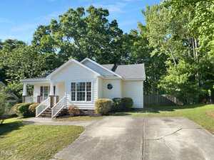 Single-story white house with a front porch, large windows, and gray roof. Surrounded by trees, it has a concrete driveway and a grassy yard. A wooden fence is visible along the right and back sides.