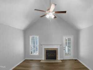 A living room with light gray walls, a vaulted ceiling, a ceiling fan with lights, a fireplace with a white mantel, a window on the left, a glass door on the right, and wood flooring.
