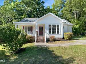 Charming single-story white house at 2747 Michelle Court, Creedmoor, with a covered front porch, red door, large windows, brick steps, and landscaped shrubs nestled among green trees on a sunny day.