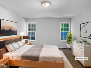 A bedroom with a wooden bed, white and gray bedding, two windows, a large dresser with plants and decor, framed art on two walls, and neutral-colored walls and carpet. The image is virtually staged.