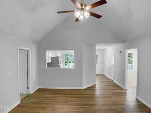 A room with vaulted white ceilings, wood flooring, a ceiling fan with lights, and an open window cutout to the kitchen. There are doorways leading to other rooms and natural light coming through the windows and doors.