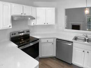 A modern kitchen with white cabinets, stainless steel appliances including a stove and dishwasher, a double sink, and wood flooring. A pass-through window reveals part of another room with a fireplace.