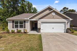 Charming single-story suburban house at 1574 Dorsett Lane, Creedmoor, featuring beige siding, brown accent shingles, white trim, and a two-car garage. A concrete driveway leads to the entrance with simple landscaping and trees in the background.