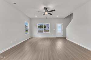 A spacious, empty living room with light wood flooring, white walls, a ceiling fan with lights, two windows, a white door with glass panel, and a staircase leading upward.