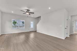 A spacious, empty living room with light wood flooring, white walls, a ceiling fan with light, recessed lighting, a window, a door leading outside, and a staircase to the upper floor.