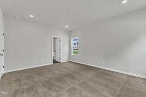 Empty room with beige carpet, white walls, a single window, and a doorway leading to a bathroom. Recessed ceiling lights are on, and there is natural light coming through the window.