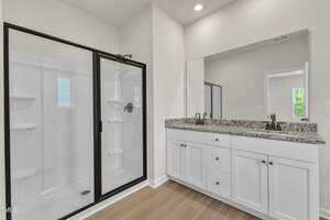 A bathroom with a glass-enclosed shower, white cabinets with granite countertop, double sinks with faucets, large mirror above the vanity, light walls, and wood-look flooring.