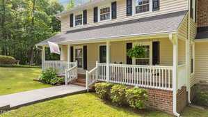 A two-story house with beige siding, black shutters, and a covered front porch. The porch has white railings, two steps, potted plants, and a hanging fern. A flag hangs near the entrance. The yard is grassy and well-kept.