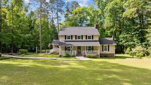 Two-story house with light-colored siding, dark shutters, and a covered front porch sits on a large, grassy lawn with a curved driveway, surrounded by tall trees under a bright sky.
