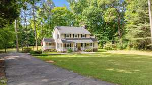 A two-story yellow house with white trim and a covered front porch sits on a large green lawn, surrounded by trees. A long driveway leads up to the house. The setting appears to be quiet and wooded.
